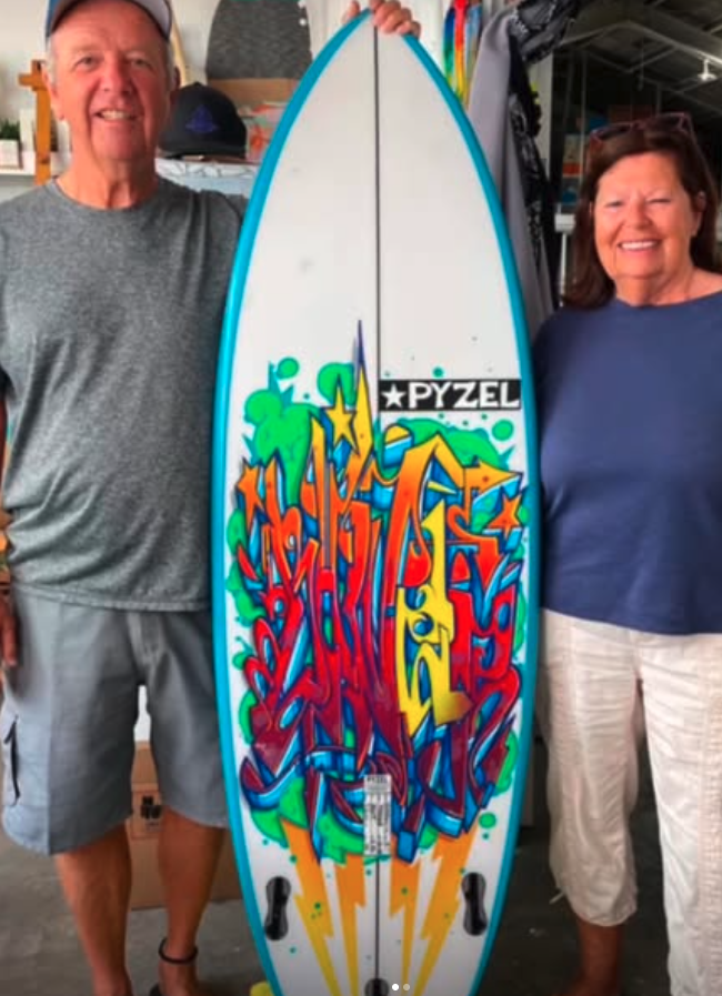 Vinny and a woman holding a surfboard painted with bold, colorful graffiti